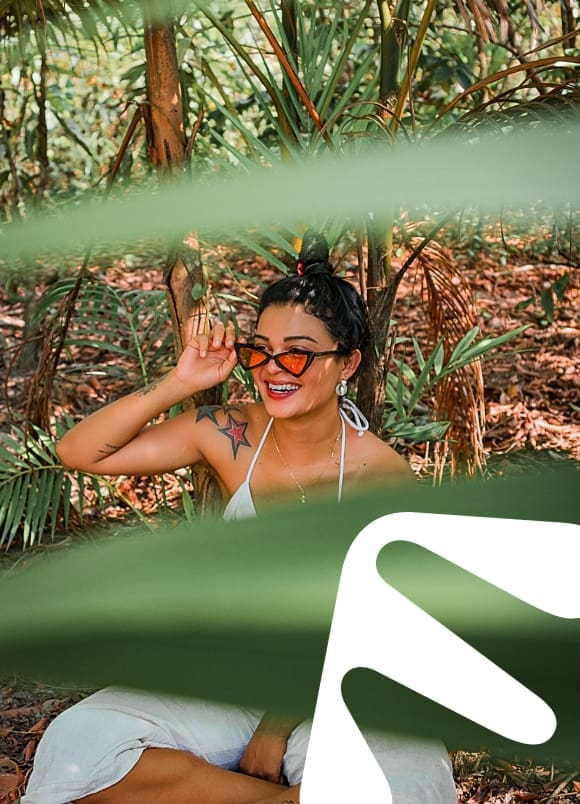 Woman holding sunglasses and smiling in a tropical setting.